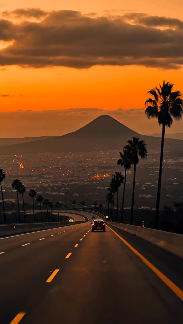 Red car driving on a palm-lined road toward a mountain at sunset.