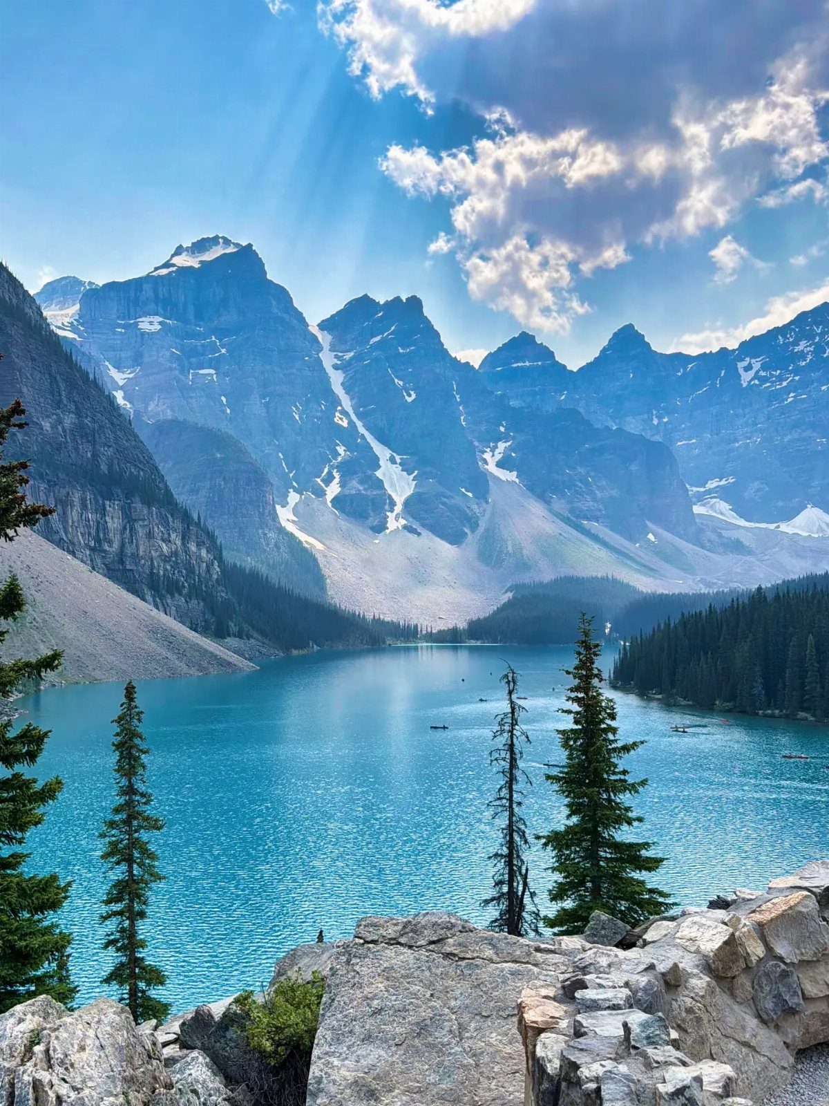 Turquoise lake surrounded by jagged snow-capped mountains and evergreen trees under sunbeams.