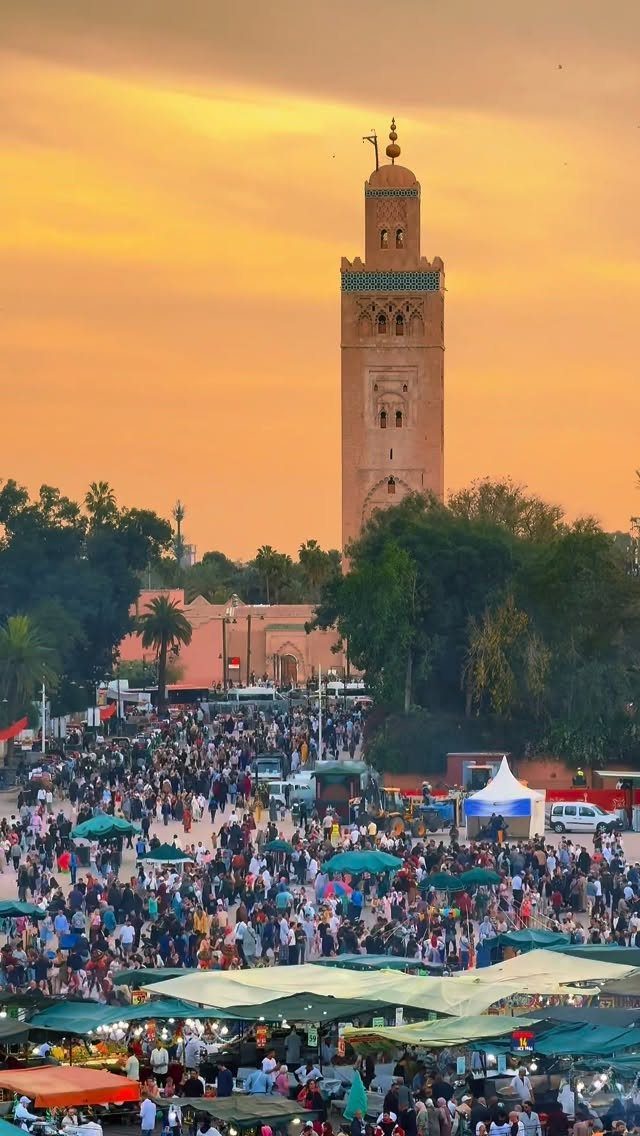 Crowded outdoor market with green umbrellas and a tall minaret under an orange sky.