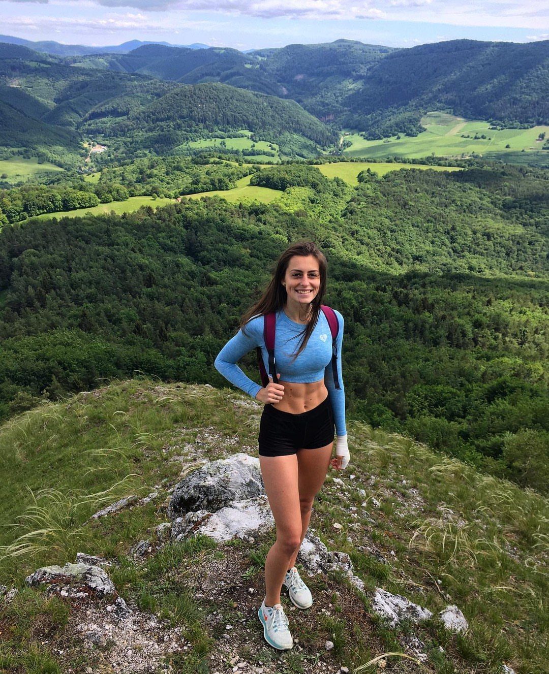 Woman with a backpack standing on a mountain peak overlooking a green valley.