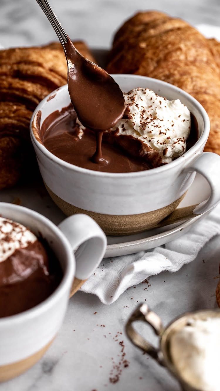 Two mugs of thick hot chocolate with whipped cream and golden croissants.