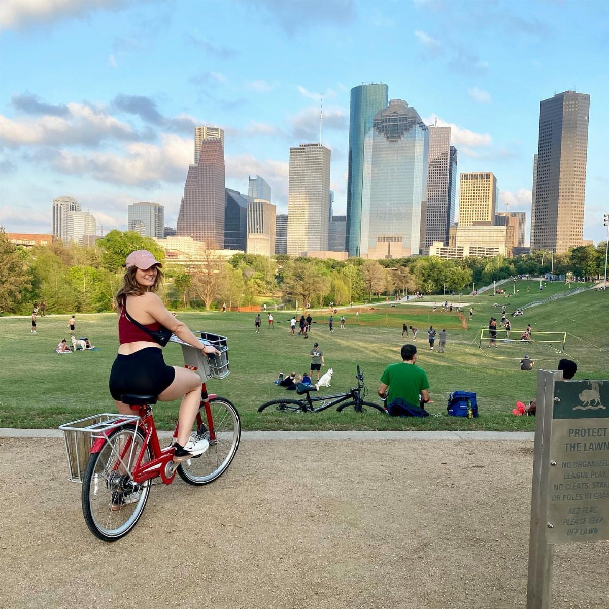 Woman riding a red bicycle on a path with a city skyline background.