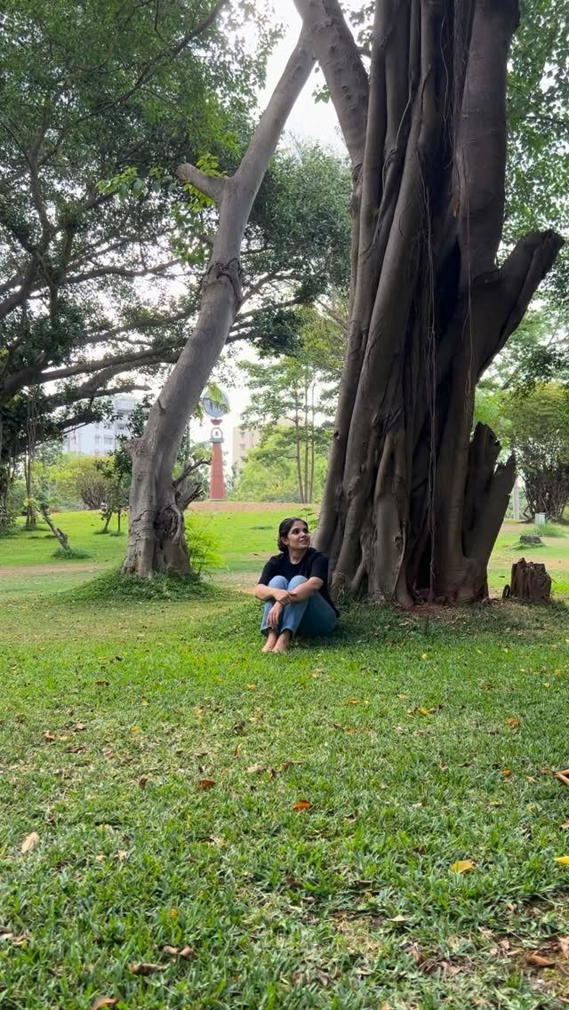Woman sitting on green grass beneath a large tree in a public park.
