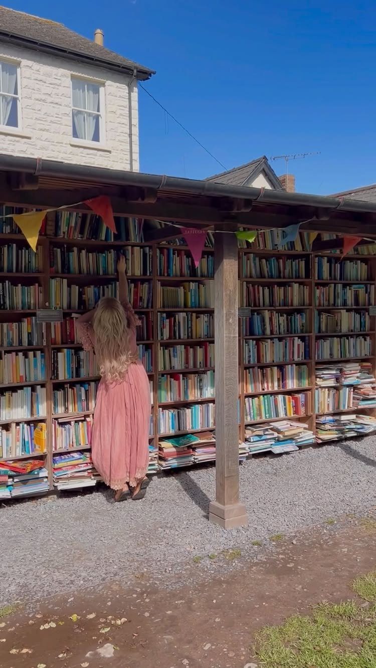 Woman in pink dress browsing a large outdoor bookshelf under a wooden canopy.