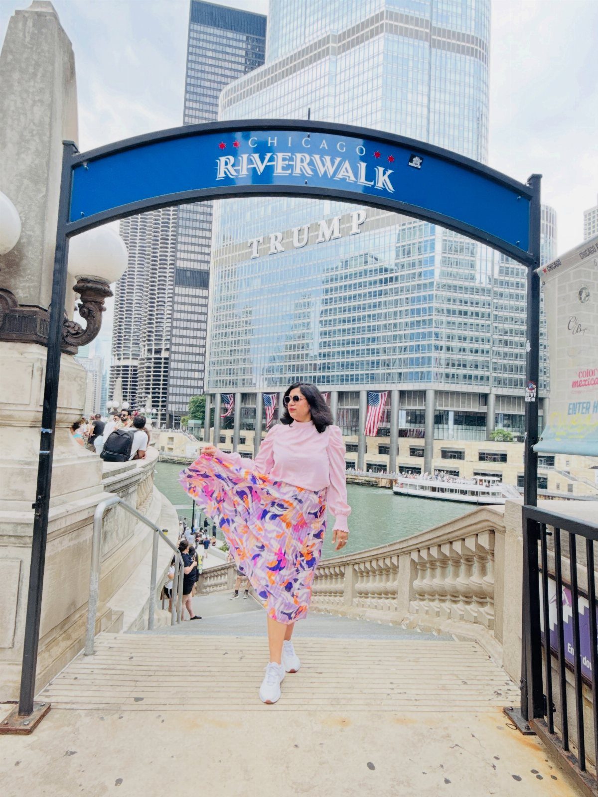Woman in colorful skirt walking under a Chicago Riverwalk sign with skyscrapers behind.