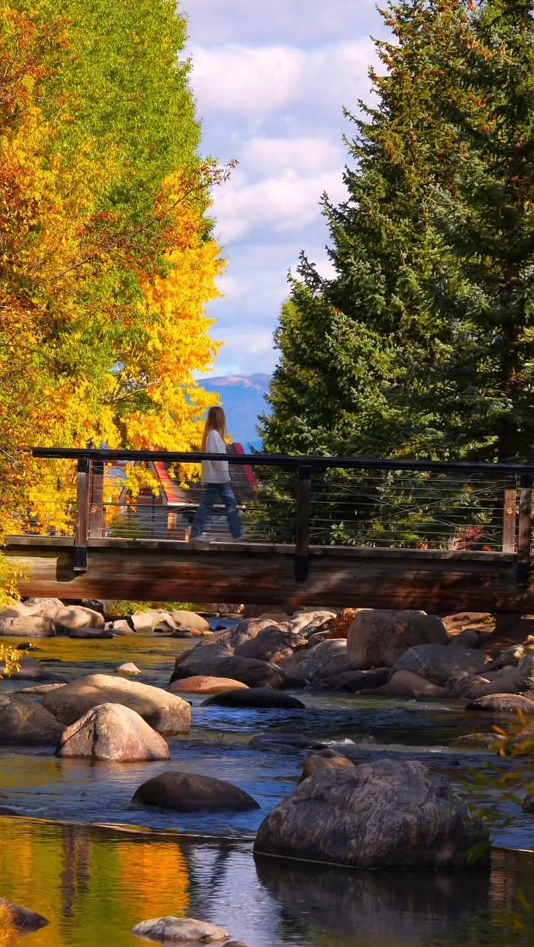 Person walking across a wooden bridge over a rocky stream with autumn foliage.