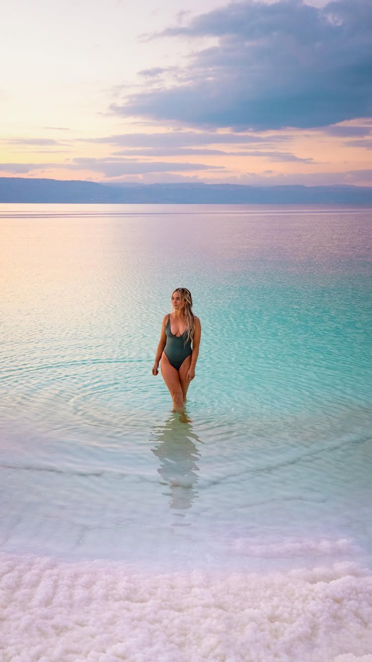 26 Pretty Places Around the World, From Fjords to Falls 15 Woman in swimsuit standing in turquoise Dead Sea water with white salt crystals.