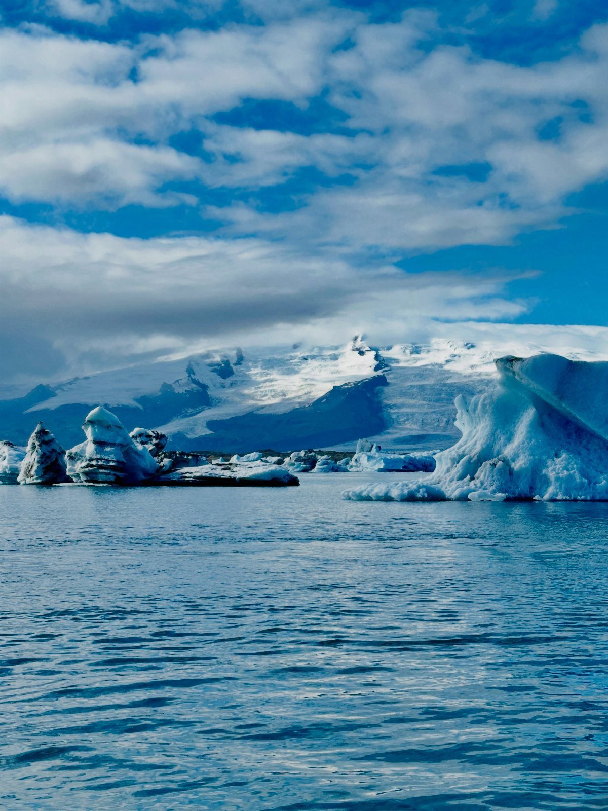 26 Pretty Places Around the World, From Fjords to Falls 22 Glacial lagoon with floating icebergs and snow-capped mountains under a cloudy blue sky.