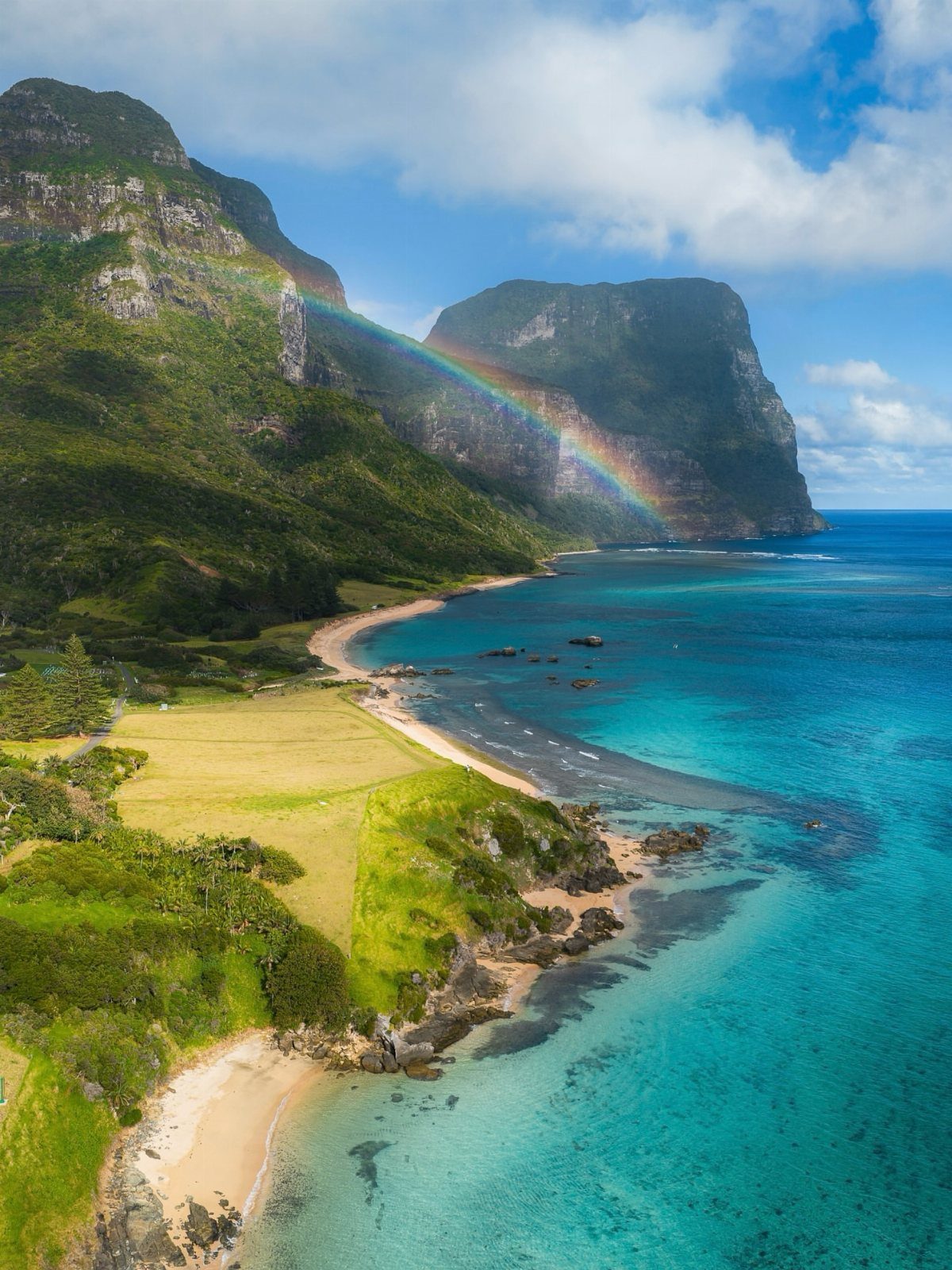 26 Pretty Places Around the World, From Fjords to Falls 10 Turquoise ocean and green mountains with a rainbow over a sandy beach coastline.