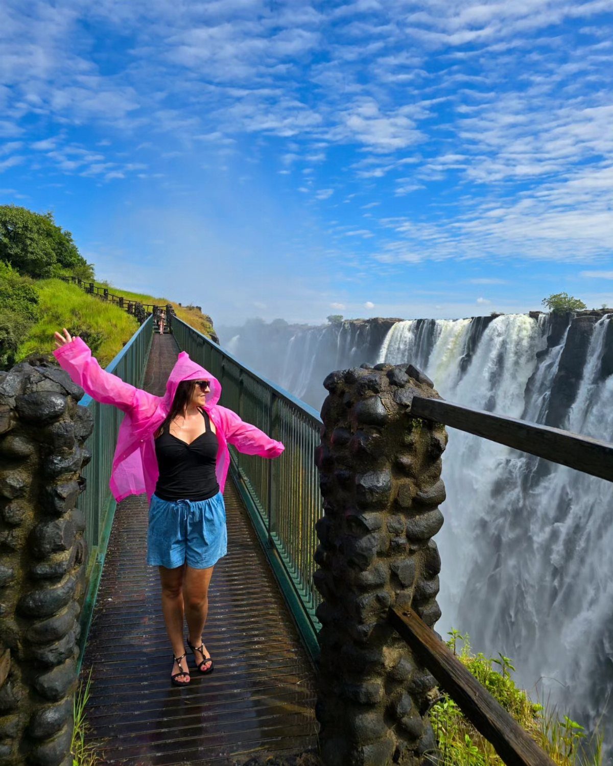 26 Pretty Places Around the World, From Fjords to Falls 12 Woman in pink raincoat standing on a walkway overlooking Victoria Falls waterfall.