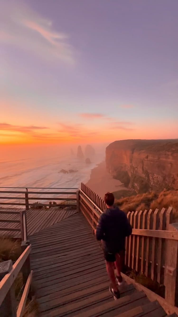 14 Pretty Places in Australia, from Reefs to Wine Regions 7 Person walking on a boardwalk overlooking the 12 Apostles limestone stacks at sunset.
