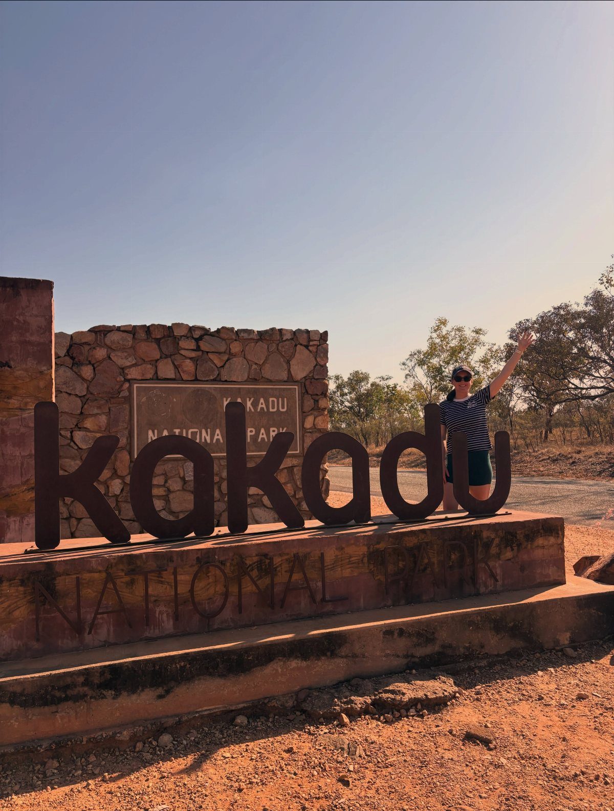 14 Pretty Places in Australia, from Reefs to Wine Regions 3 Woman waving beside the large Kakadu National Park sign in a red desert landscape.