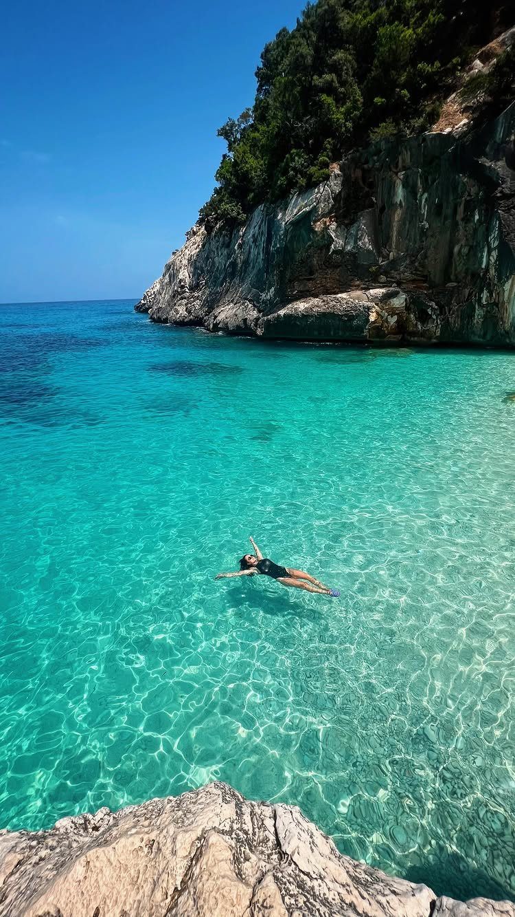 13 Pretty Places in Italy Worth Visiting in 2026 11 Person floating in turquoise water next to a rocky cliff and green trees.