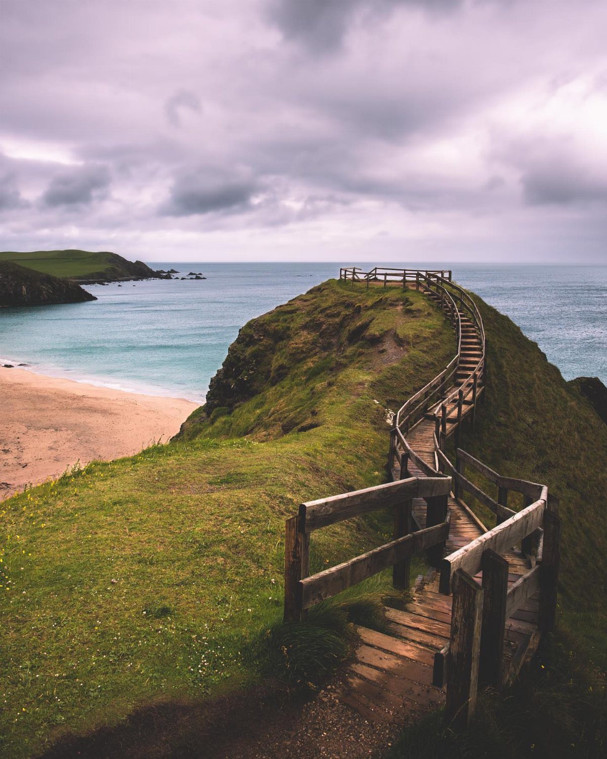 Wooden boardwalk winding along a grassy cliff overlooking a beach and ocean.