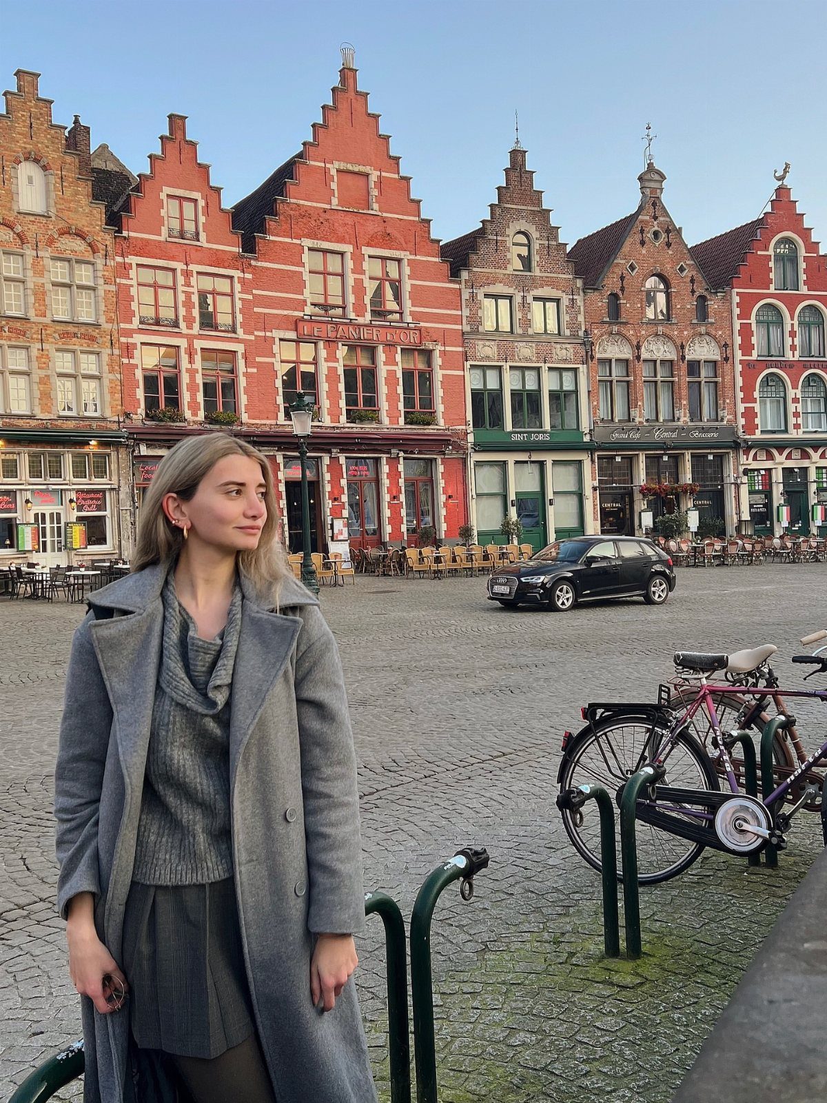 Woman in grey coat standing before historic stepped-gable buildings in Bruges, Belgium.