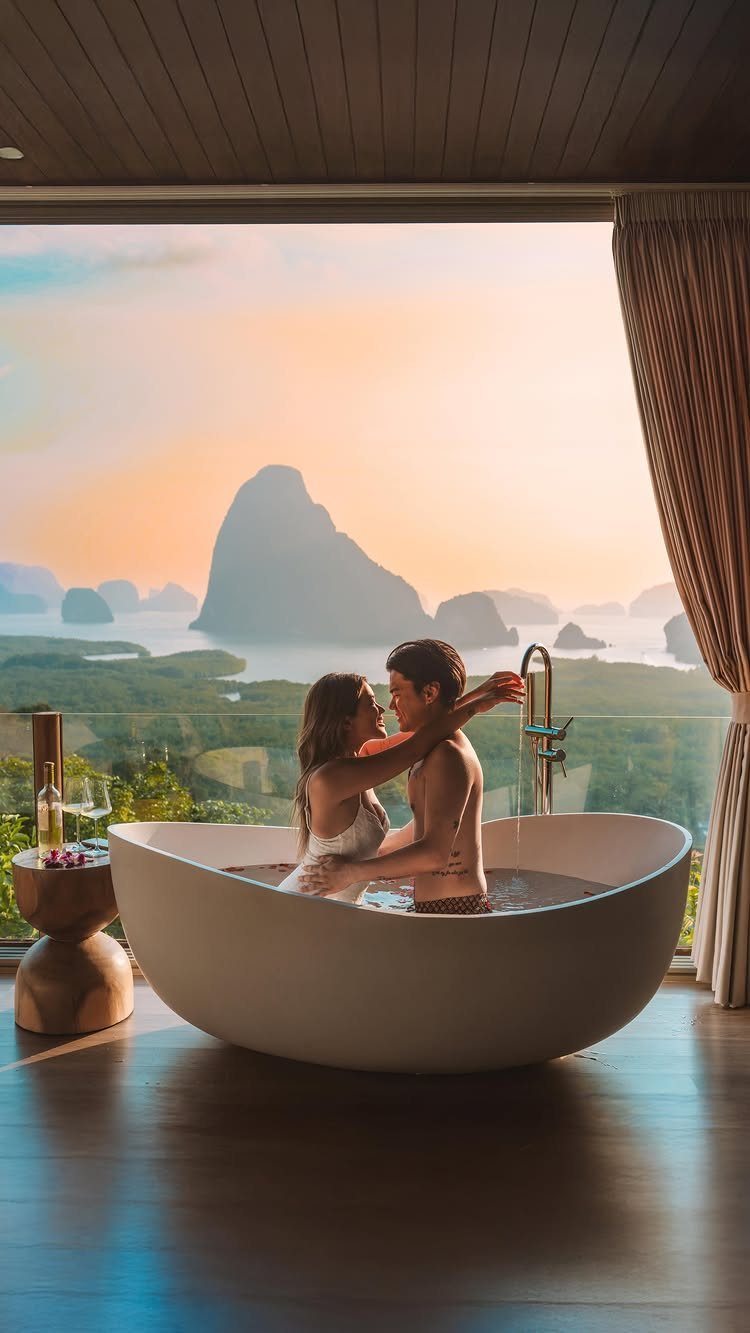 Couple embracing in a bathtub overlooking tropical limestone karsts and a sunset sea.