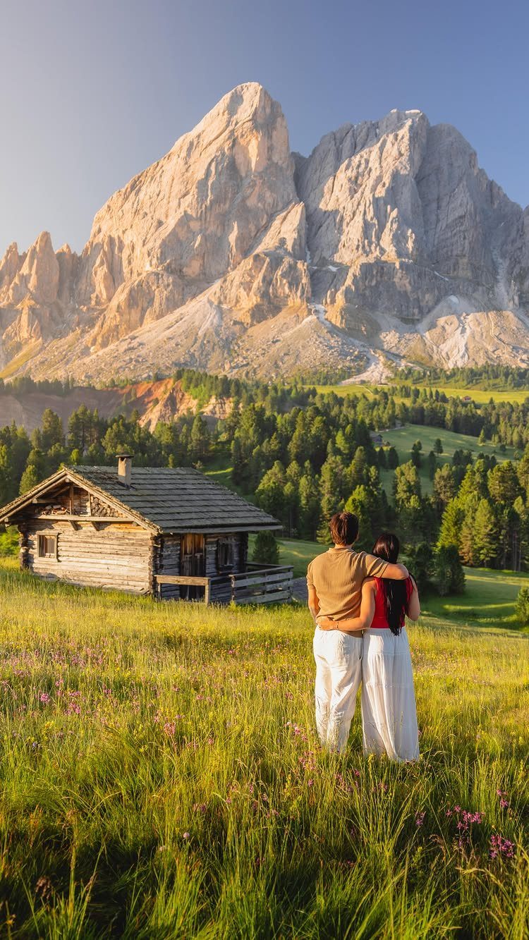 Couple embracing in a mountain meadow with a rustic cabin and jagged peaks.