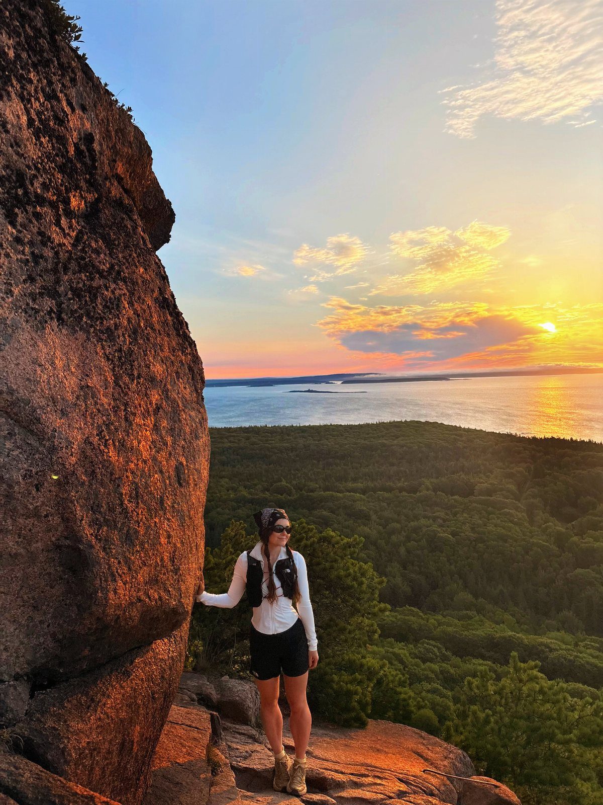 7 Sights to See in the US, From Acadia to Old Idaho 1 Woman standing on a rocky cliff overlooking a forest and ocean at sunset.