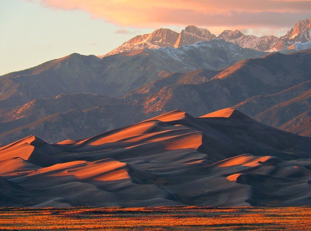 7 Sights to See in the US, From Acadia to Old Idaho 2 Orange sand dunes stretching toward snow-capped mountains under a sunset sky.