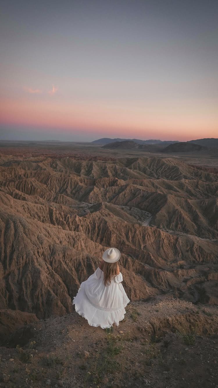 Woman in white dress overlooking vast, eroded brown badlands hills at sunset.