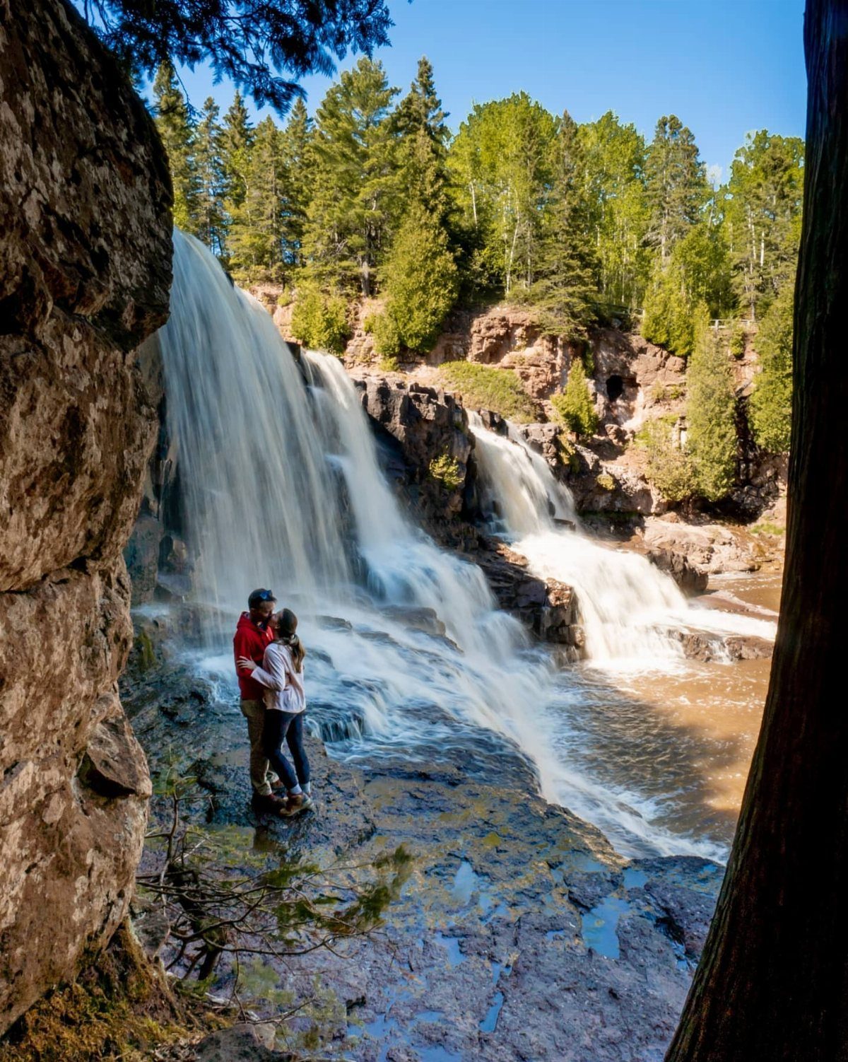 Couple standing on rocks before a large cascading waterfall surrounded by green forest.