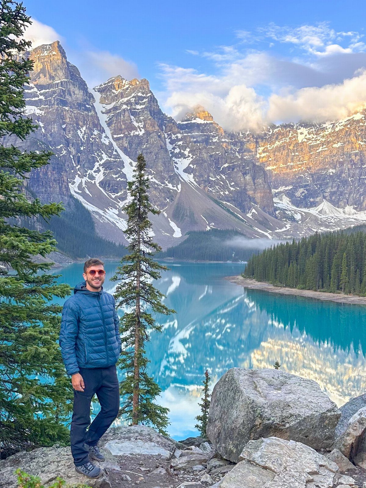 Man standing on rocky cliff overlooking a turquoise lake and snowy mountains.