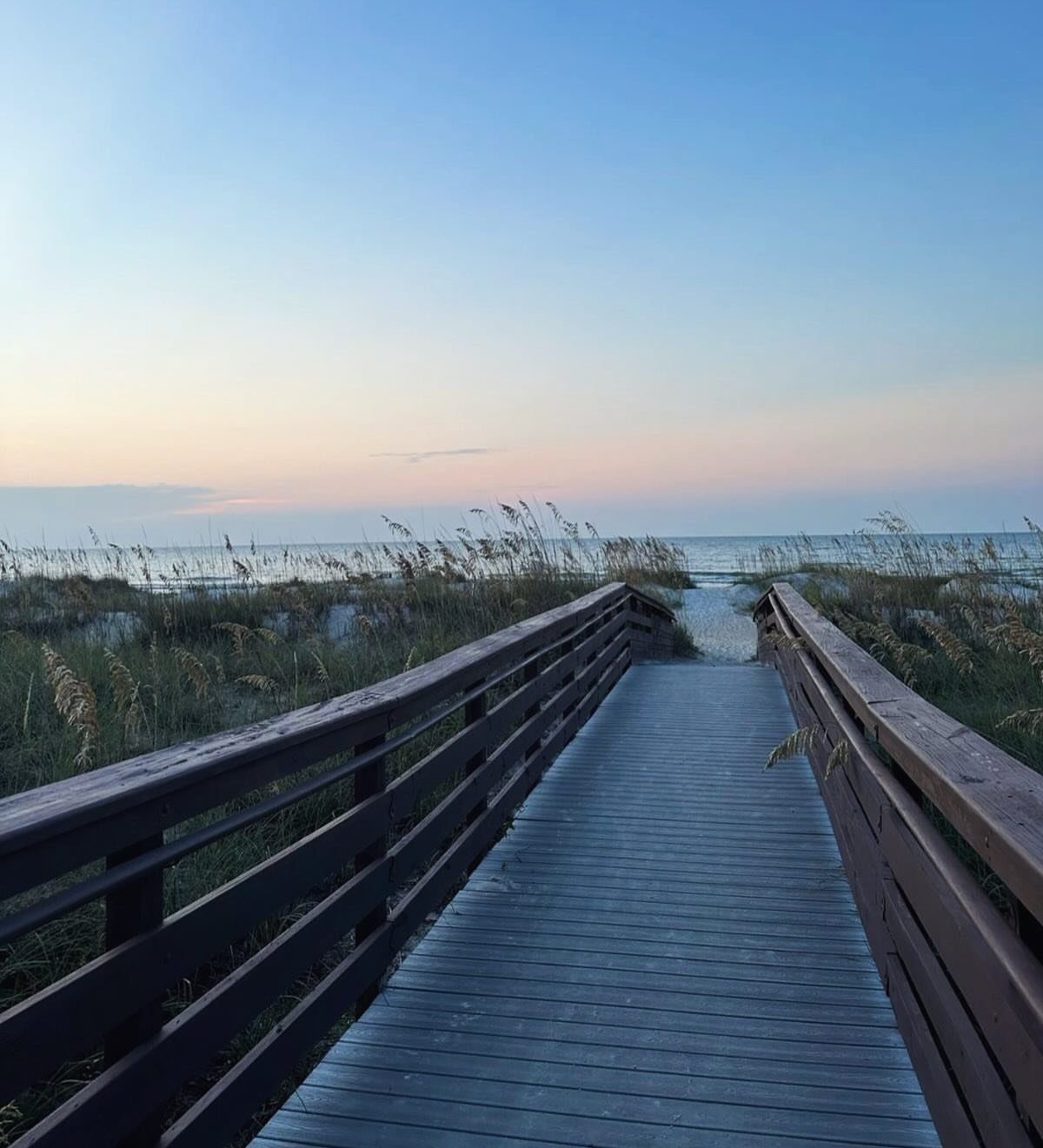 Wooden boardwalk leading through beach grass toward the ocean at twilight.