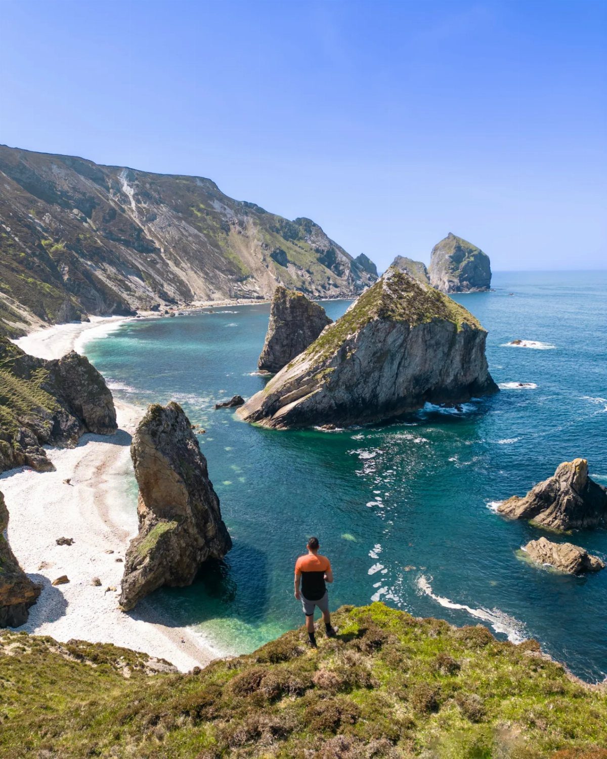 Man standing on a grassy cliff overlooking a turquoise bay with rocky islands.