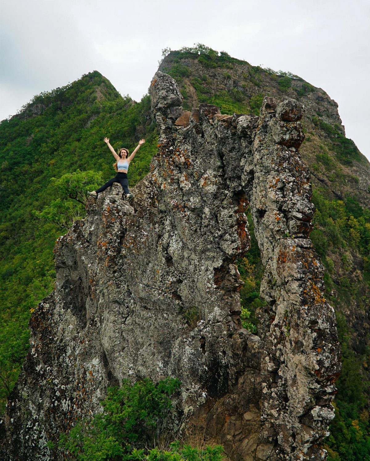 Woman standing with arms raised on a high, jagged rock peak among green mountains.