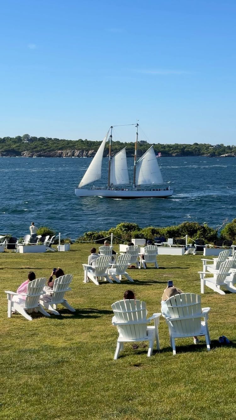 White sailboat on blue water viewed from a lawn with white Adirondack chairs.