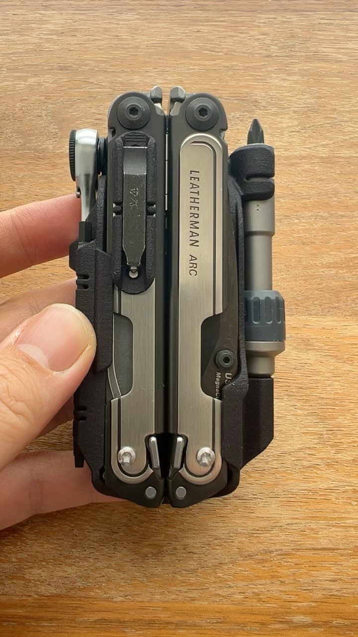 Leatherman Arc multi-tool held in a hand over a wooden background.