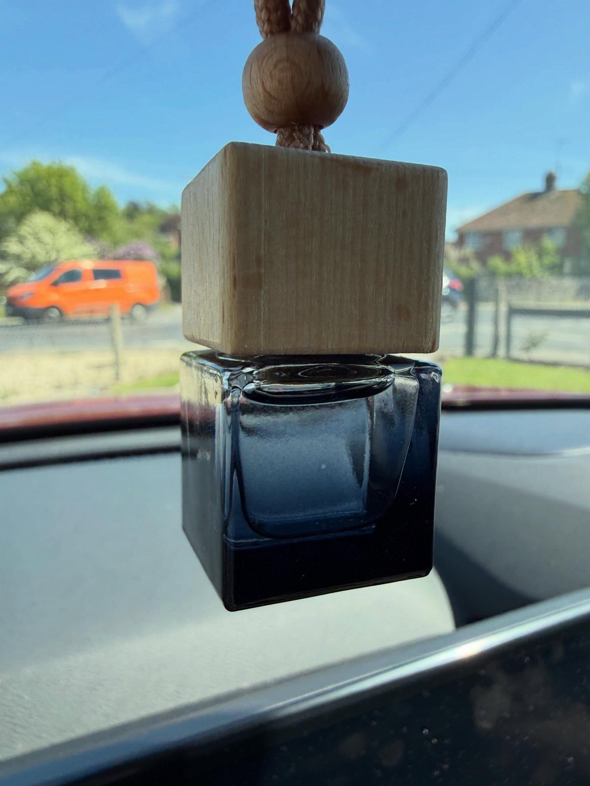 Square car air freshener with wooden top and glass bottle hanging in vehicle.