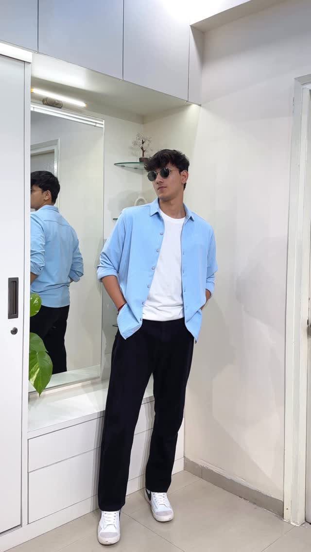 Man wearing a light blue shirt layered over a white t-shirt and black trousers.