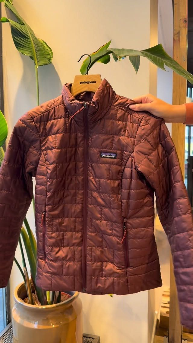 Maroon quilted lightweight jacket hanging on a wooden hanger next to a green plant.