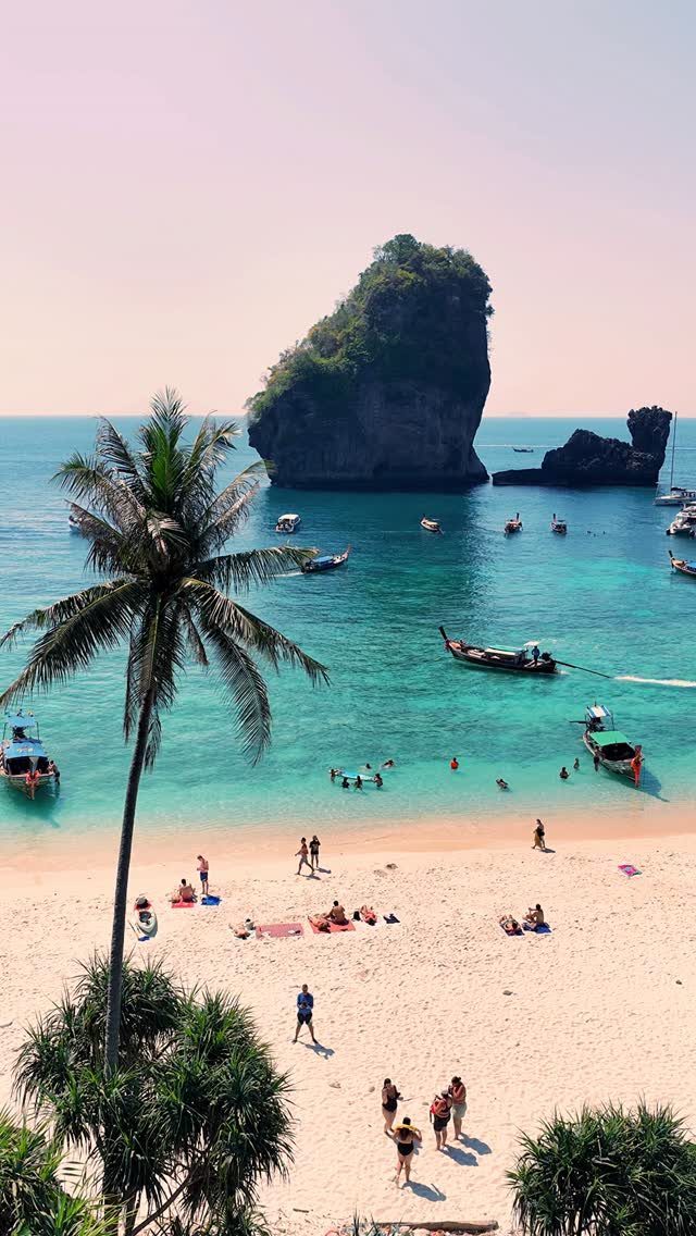 Turquoise bay with limestone cliffs, long-tail boats, and people on a white beach.