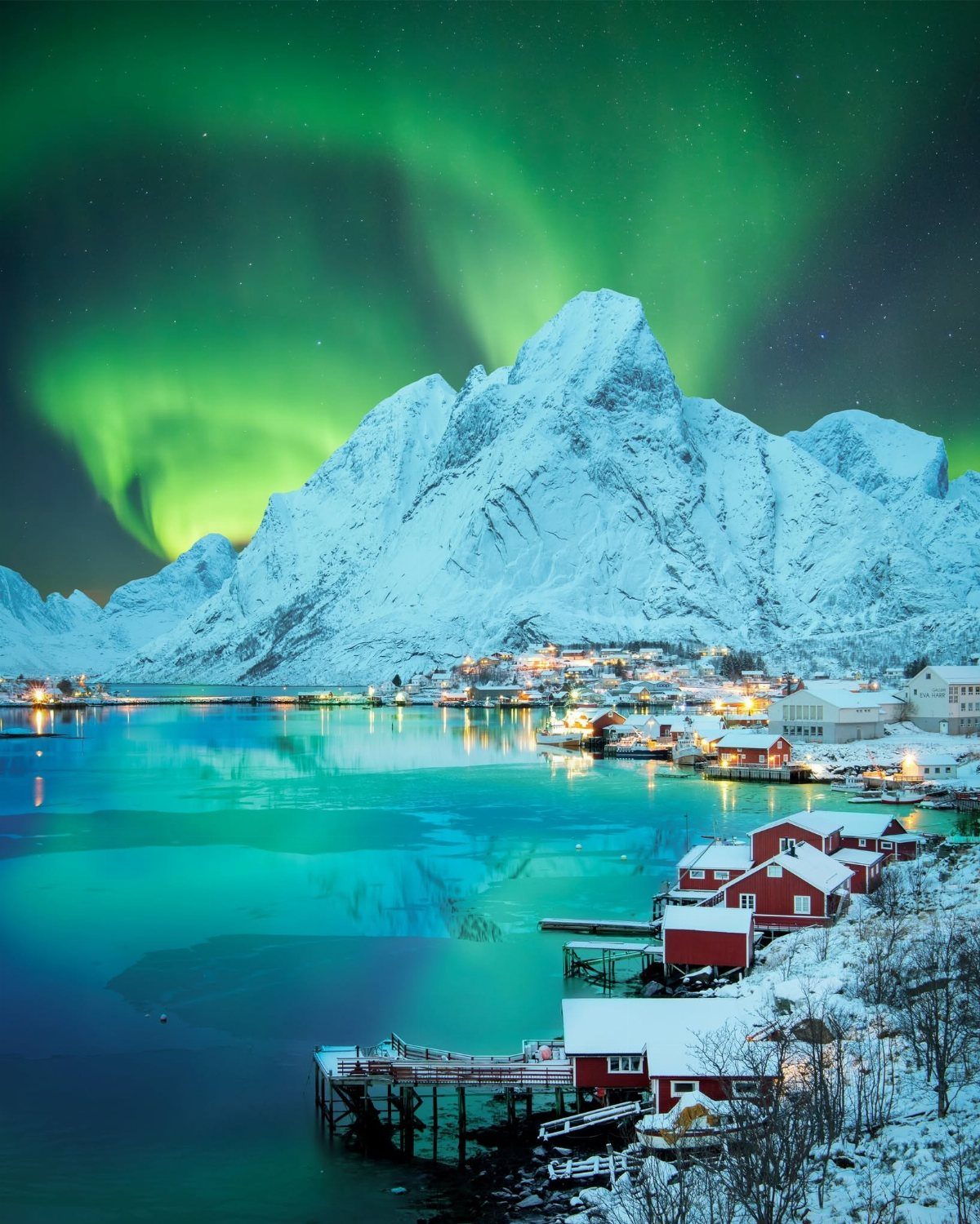 Green northern lights over snowy mountains and a red fishing village by the water