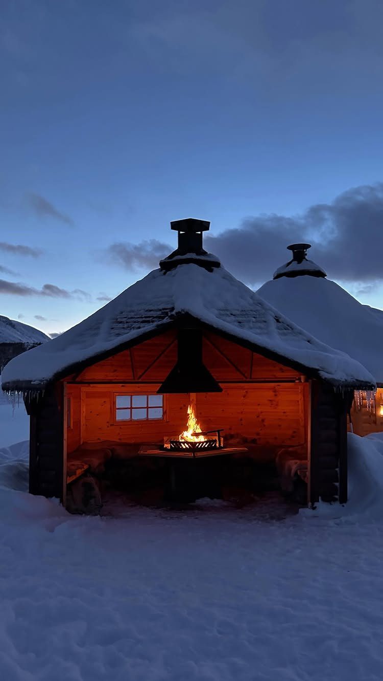 Snow-covered wooden shelter with a warm campfire glowing inside at twilight.