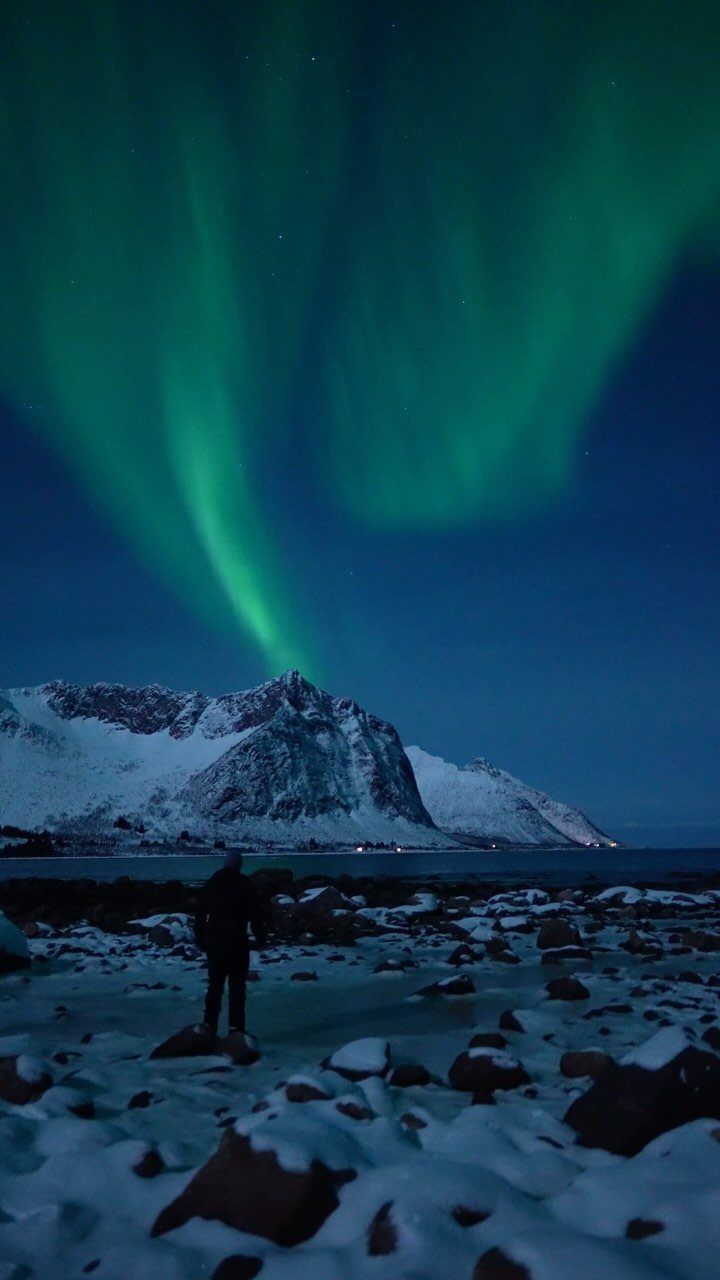Green aurora borealis dancing over a snowy mountain and rocky coastline at night
