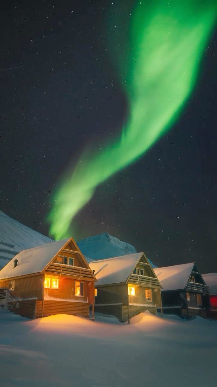 Wooden cabins in snow under a bright green aurora borealis night sky