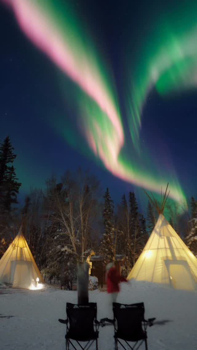 Aurora borealis swirling over snowy ground with illuminated tipis and folding chairs.
