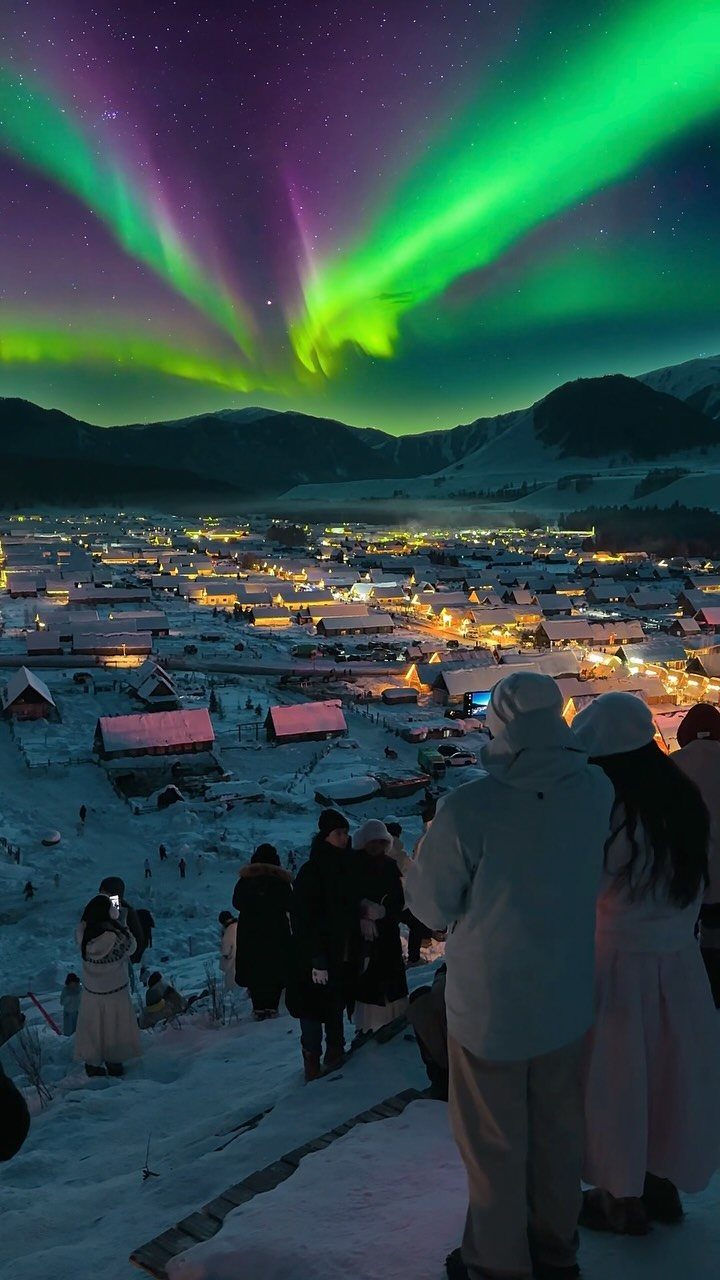 Aurora borealis glowing green and purple over a snowy village with people watching.