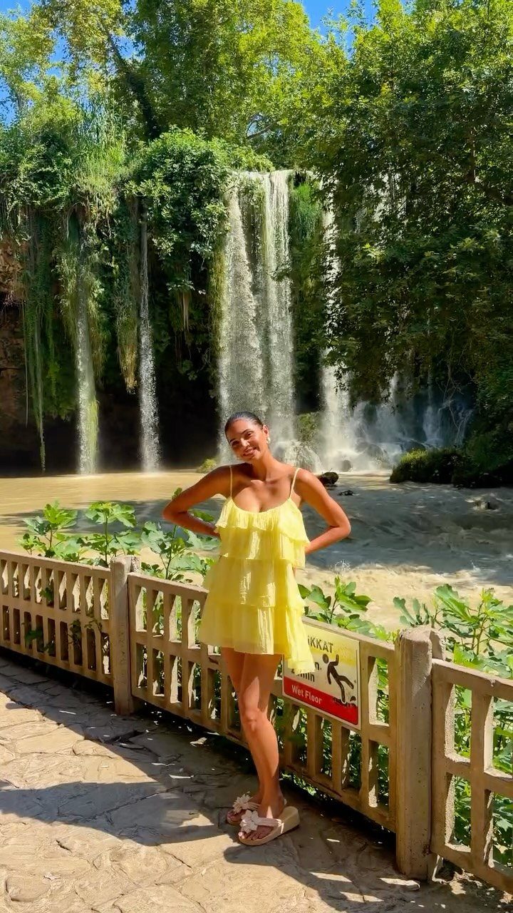 Woman in yellow dress posing before a tall waterfall and green forest