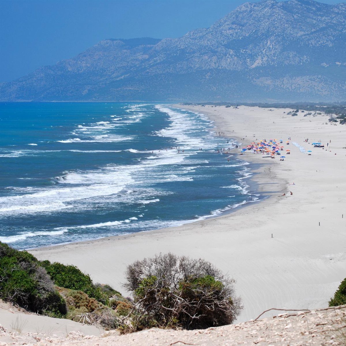 White sandy beach with turquoise water and mountains under a clear blue sky.
