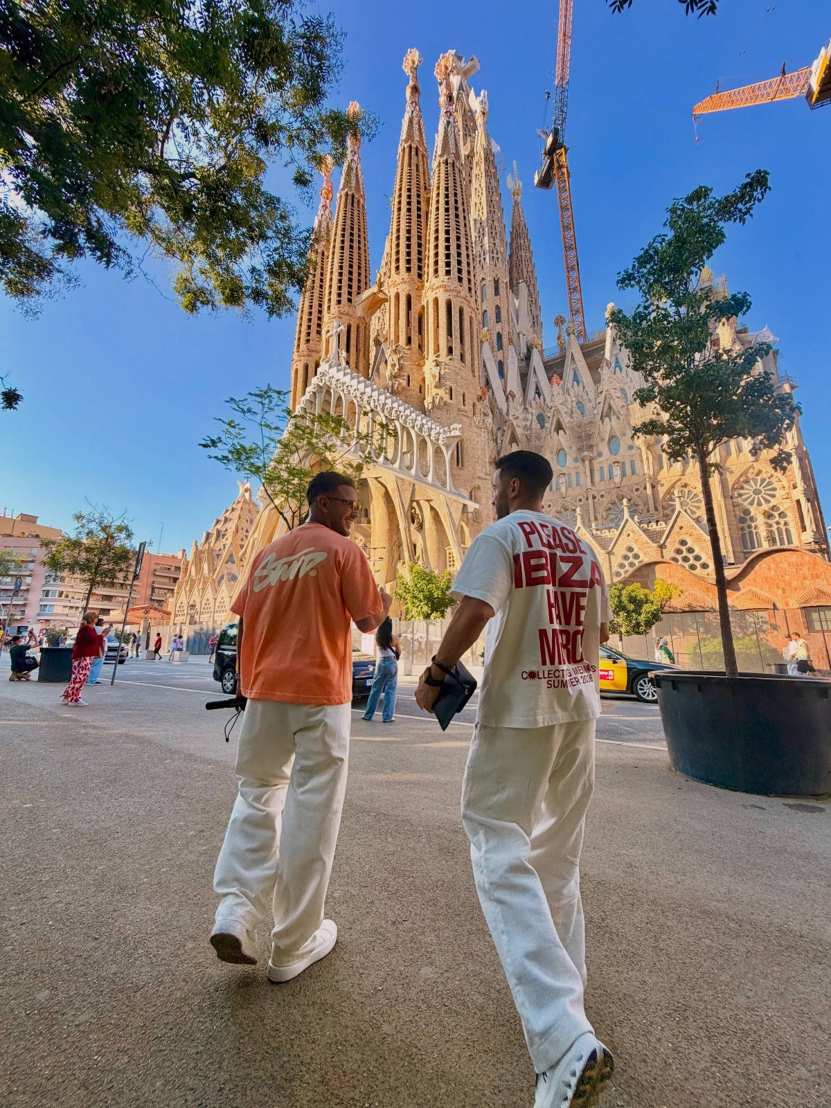 12 Vacation Ideas with Friends, from Cruises to Cancun 5 Two men walking toward the Sagrada Familia basilica under a clear blue sky.