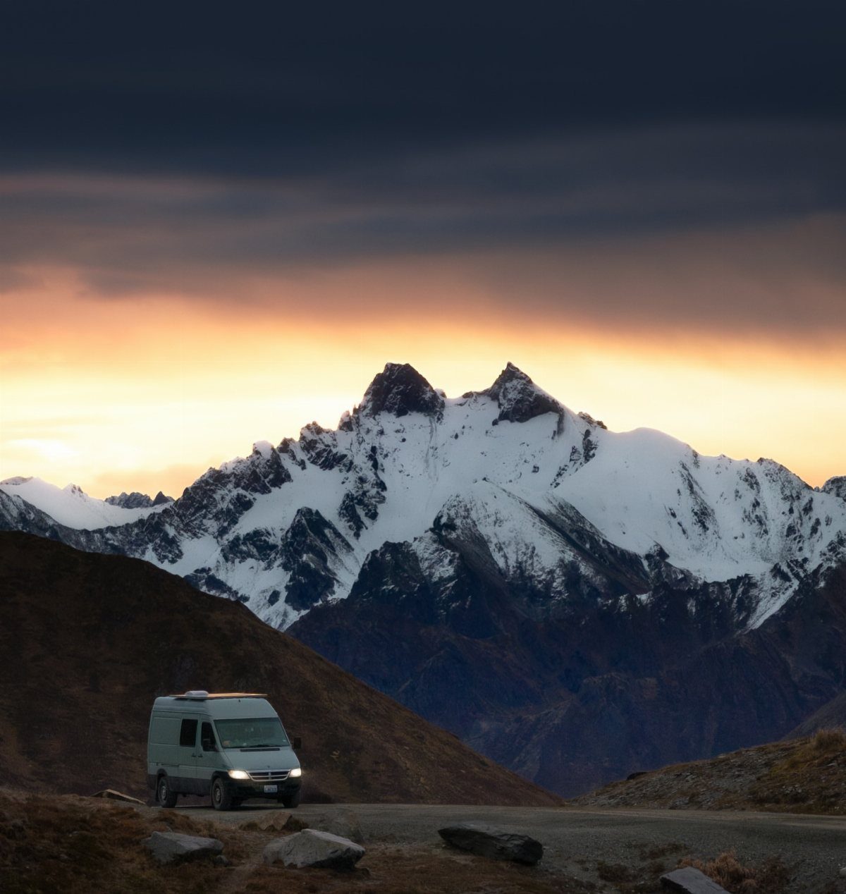 White camper van parked before massive snow-capped mountains under a dramatic sunset sky.