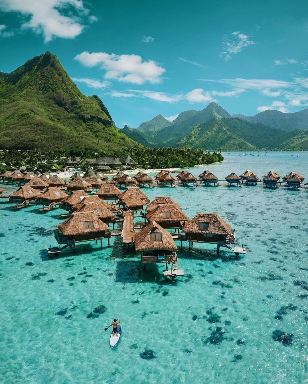 Overwater bungalows with thatched roofs in turquoise water against lush green mountains.