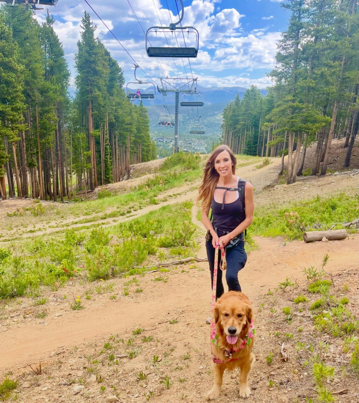 Woman and golden retriever on a mountain trail with ski lifts and pines.