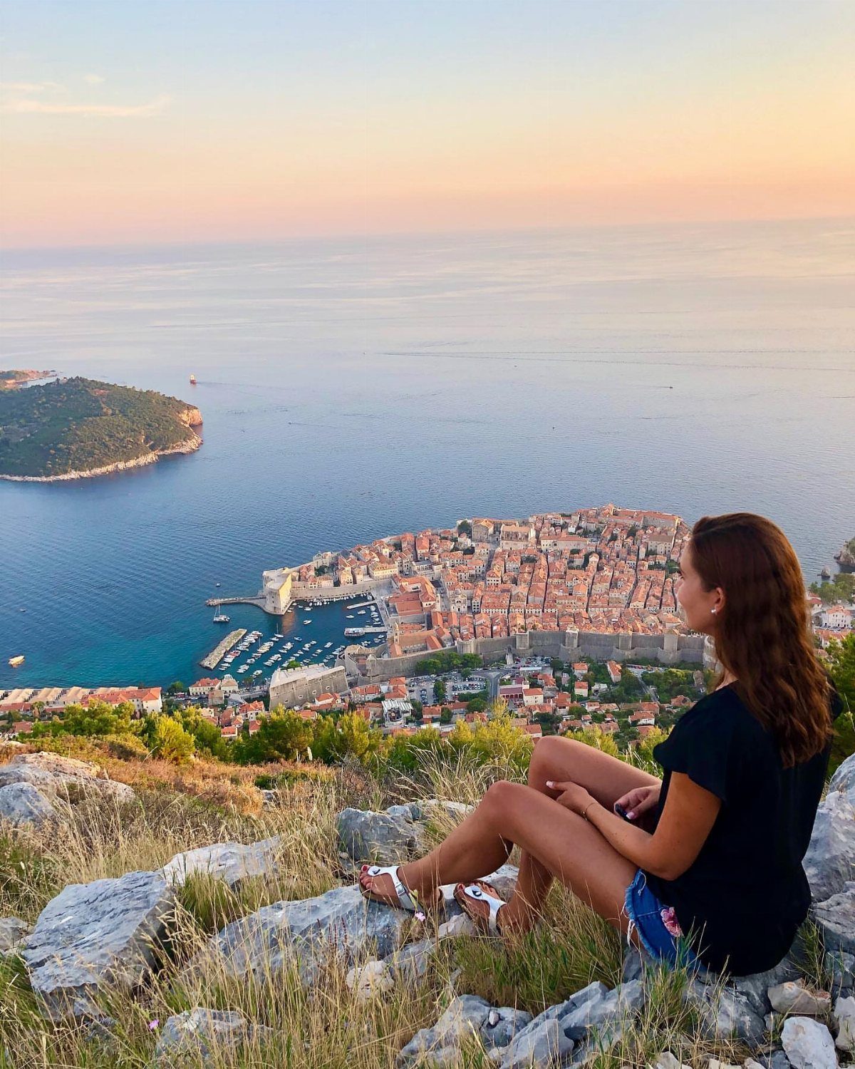 Woman sitting on a rocky cliff overlooking a historic coastal city and harbor.