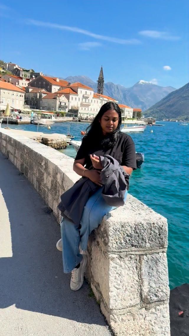 Woman sitting on a stone wall overlooking a coastal town with orange roofs.