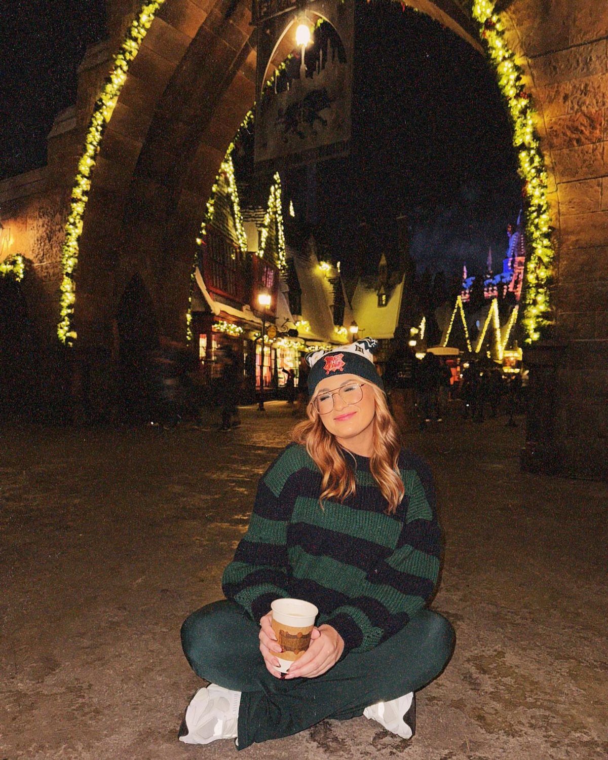 Woman in winter clothes sitting under a lit archway in a festive village